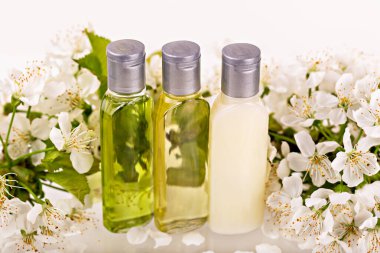 Hassas beyaz çiçeklerin arasına yerleştirilmiş gümüş kapaklı üç doğal kozmetik şişe. Şeffaf ve kremsi dokular organik cilt ürünleri olduğunu gösteriyor. Güzellik, sağlık ve kaplıca kavramı