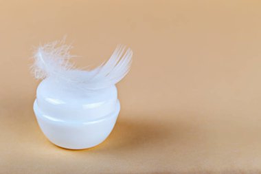Modelleme. Yumuşak pastel arka planda beyaz tüyle süslenmiş krema kavanozu. Krem cilt yumuşaklığı konseptini taşır. Kozmetik krem kavanozu güzellik rutinini ve hassas cilt bakımını ön plana çıkarıyor.