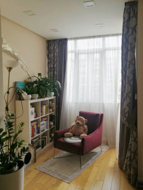 Kyiv, Ukrayna - 26 Mart 2025: Kırmızı koltuklu rahat okuma köşesi, oyuncak ayı, kitap rafları, ev bitkileri ve parlak iç kısımdaki perdeler. Yumuşak ışıklandırma huzurlu bir ev atmosferi yaratır. Dikey