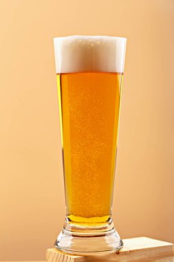 Bir bardak köpüklü bira, ahşap bloğun üzerine bej arka plana yerleştirilmiş. Bira içeceği, altın bira ve içecek yaşam tarzı kavramı. Bira lezzeti ve gelişigüzel anları simgeler.