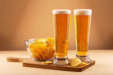 İki bardak köpüklü soğuk altın bira ve tahta tahtada servis edilen patates cipsi. Bira çerezi, bar yemeği ve rahatlama zamanı. Bira ve cips rahat olduğunu gösterir.