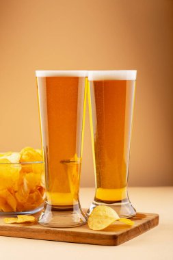 İki bardak köpüklü altın bira ve bir kase patates cipsi tahtada sıcak arka planda. Bira ve aperatif kavramı, bar ve günlük içecek. Uçakta dikey bira ve patates kızartması var.