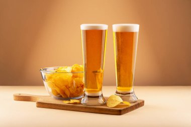 Cam kasede çıtır patates cipsi ve sıcak arka planda iki soğuk bira. Bar atıştırmalığı, sosyal içecek ve sıradan bira eşleşmesi kavramı. Bira ve cips rahatlığı ve tadı yansıtır.