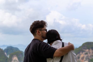 Romantik çift, çarpıcı dağlar ve dramatik gökyüzü ile çevrili, manzaralı bir bakış açısıyla samimi bir kucaklaşmayı paylaşıyor, sevgi ve sükuneti çağrıştırıyor. Manzaralı manzaraya sarılan çift