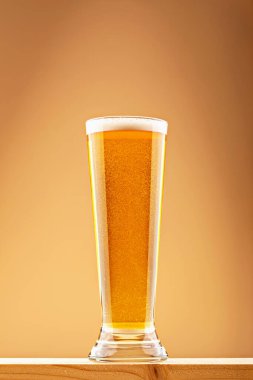 Sıcak ortam ışığında, ahşap bir kalas üzerinde hafif köpüklü, yarım litre parlak altın bira. Bira ferahlatıcı ve bira berraklığı kavramı. Tahta yüzeyinde köpük olan uzun altın bira bardağı.