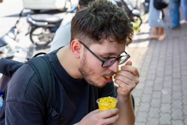 Sırt çantalı gözlüklü genç adam, arka planda bulanık figürler ve araçlarla çevrili kentsel çevre ile çevrili kaldırımda otururken bir fincan mısırın tadını çıkarıyor. Sırt çantalı gezgin.