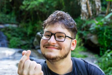 Gözlüklü genç adam gülümsüyor ve elleriyle kalp hareketi yapıyor, canlı yeşillik ve doğal elementlerle çevrili, neşeli ve dost canlısı atmosfer yaratıyor. Sırt çantalı gezgin.