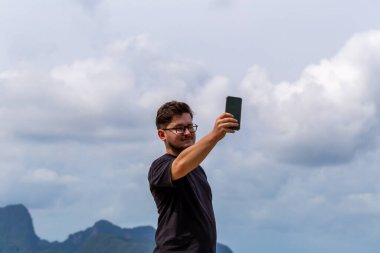 Gözlüklü genç adam açık havada dağların ve bulutların manzaralı zeminine karşı selfie çekiyor. Doğadaki neşe ve macera anını yakalıyor. Erkek şahıs dışarıda selfie çekiyor.