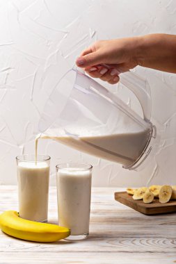 Muzlu milkshake. El, şeffaf sürahiden, taze muz ve dilimlenmiş meyveyle birlikte ahşap masada duran iki uzun bardağa kremalı muz smoothie dökerek canlandırıcı bir sahne yaratıyor.