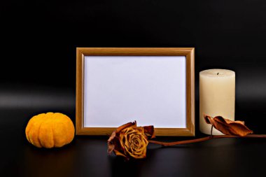 Cadılar Bayramı. Modelleme. Boş bir ahşap çerçevenin yanında canlı balkabağı, kurumuş gül ve mum içeren dekoratif bir sonbahar düzenlemesi, zengin doku ve renklerle dolu samimi bir Cadılar Bayramı atmosferi yaratıyor.