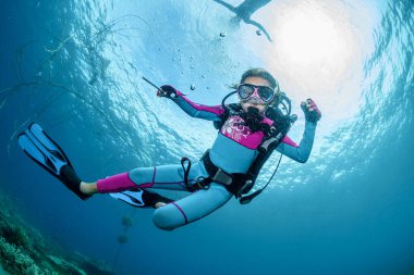 Happy little girl dives into the blue water of the ocean in scuba gear, mask and fins in the rays of sunlight, looking at the camera at the photographer. Lifestyle travel, beach adventure, summer
