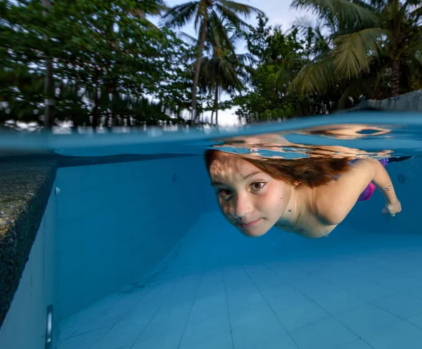 A little girl in the pool dives underwater against the backdrop of palm trees in the sunlight. Fun in the pool for children. Family vacation at the hotel. Weekend by the sea. Freediving training for