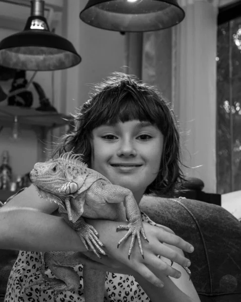 9-10 yaşlarında genç bir kız kameraya bakıp gülümsüyor ve elinde yeşil bir iguana tutuyor. İguana iguana sp., öngörünüm