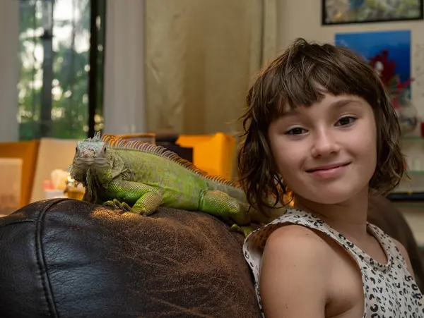 9-10 yaşlarında gülümseyen bir kız. Oturma odasında büyük bir kertenkelenin yanında oturuyor. İguana iguanası.