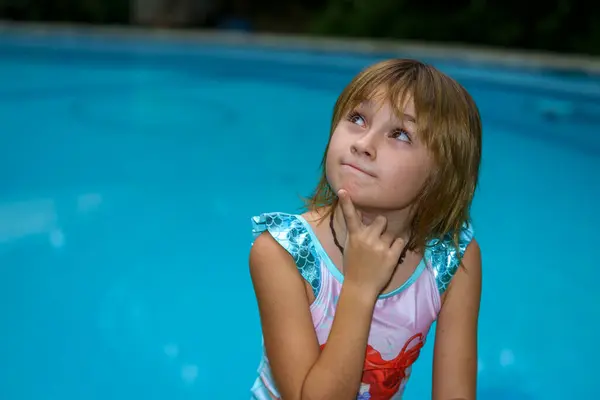 Yüzünde şüpheli ve düşünceli bir ifadeyle havuz kenarında 8-9 yaşlarında mayo giymiş bir kız..
