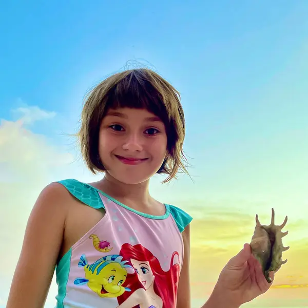 Kumsalda elinde deniz kabuğu tutan ve gülümseyen bir kız.