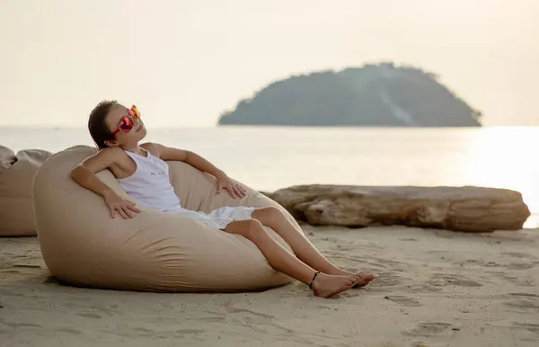Beyaz elbiseli ve güneş gözlüklü bir kız plajda dinleniyor, sandalyede oturuyor.