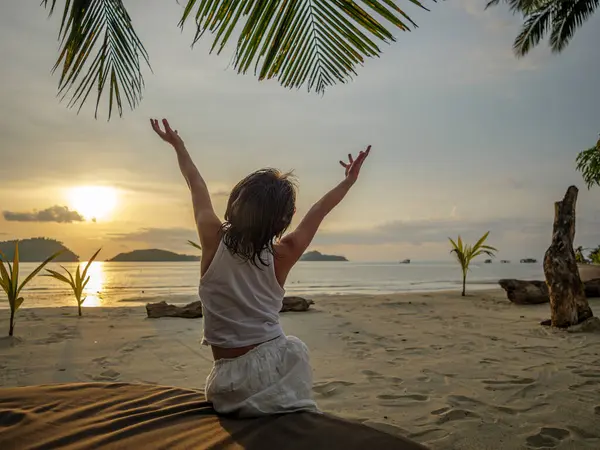 Beyaz elbiseli küçük bir kız, plajda ellerini kaldırdı palmiye ağaçlarının arka planına karşı, güzel bir gün batımı, deniz ve adalar. Arka plan