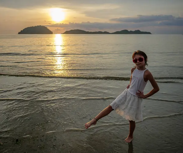 Güneş gözlüklü ve beyaz elbiseli küçük bir kız güzel bir gün batımı ve adanın arka planında duruyor.