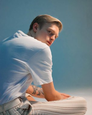 Beyaz tişörtlü genç sarışının portresi. Stüdyoda gün ışığında mavi arka planda oturuyor.