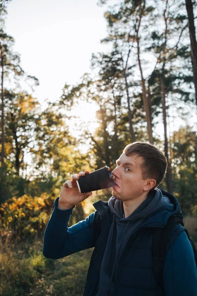一个穿着蓝色夹克的家伙在秋天的公园里喝着一杯饮料 喝着纸杯里的咖啡 — 图库照片