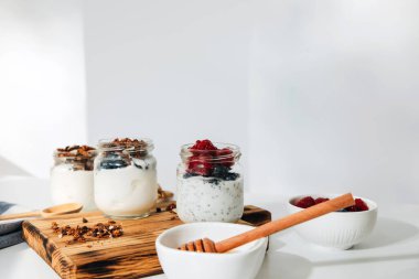 Yaban mersini, ahududu ve chia tohumlarıyla dolu cam kavanozlarda yoğurtlar ve beyaz bir zemin üzerinde tahtadan yapılmış tahıl gevreği. Sağlıklı beslenme kavramı. Ön görünüm