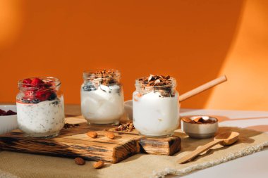 İçinde yabanmersini, ahududu ve chia tohumları olan cam kavanozlarda yoğurtlar ve turuncu bir arka planda tahtadan yapılmış tahıl gevreği. Sağlıklı beslenme kavramı. Ön görünüm