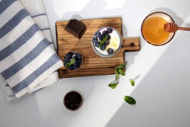 Çikolata, yaban mersini ve ballı cam kavanozda yoğurt. Beyaz arka planda, nane yapraklı ahşap bir tahtada. Düz yatıyordu