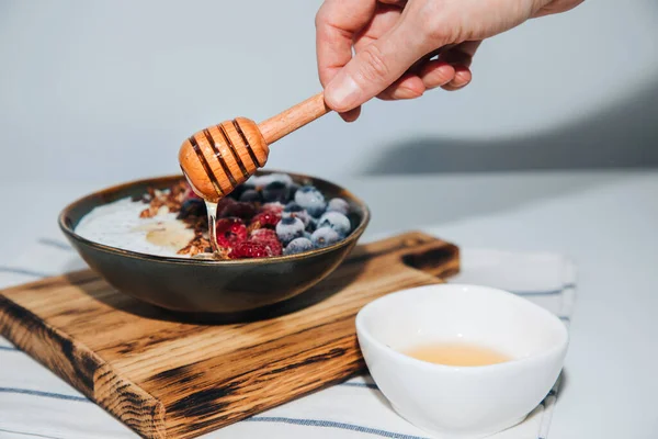 Stock image Female hand pours honey yoghurt in green plate with blueberries, raspberries, chia seeds and granola with a wooden board on a kitchen towel. The concept of healthy eating. Front view