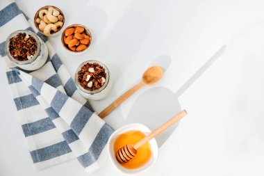 Yoğurtlar cam kavanozlarda granola, çikolata, bal ve fındıklı mutfak havlusuyla birlikte beyaz arka planda. Sağlıklı beslenme kavramı. Düz yatıyordu