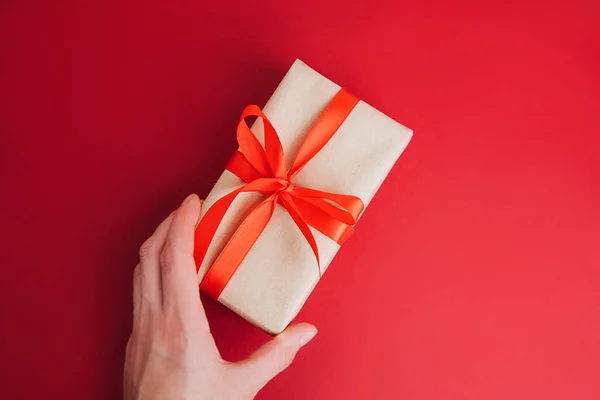 A gift wrapped in brown paper with a red ribbon on a red background is held by a female hand. Christmas gift, flat lay