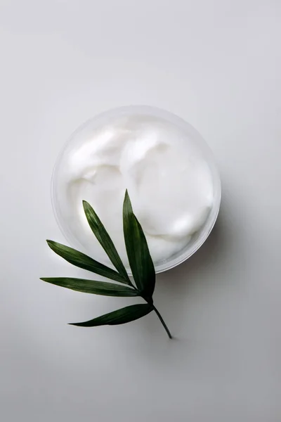 Cosmetic product of creamy texture of white color in a jar on a white background with a branch of a decorative palm tree. Flat lay