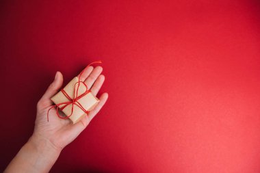 Kırmızı arka planda kırmızı kurdeleli kahverengi kağıda sarılı bir hediye bir kadın eli tarafından tutulur. Noel hediyesi, düz yat.