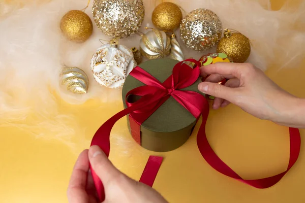 Womens hands tie a red ribbon on a green round gift. Christmas background with golden balls and angel hair. Top view