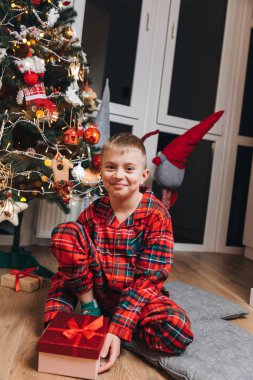 Kırmızı ve yeşil pijamalı bir çocuk elinde bir hediyeyle Noel ağacının yanında oturuyor. Mutlu Noeller ve aileniz. Ön görünüm