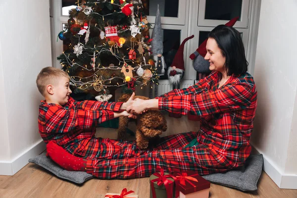 Mom and son in red pajamas and dog sit near the Christmas tree and hugging. Front view
