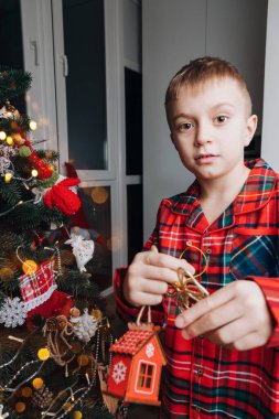 Kırmızı ve yeşil pijamalı gülümseyen bir çocuk evde bir Noel ağacı süsler. Noel ve aile. Ön görünüm