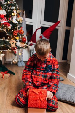 Noel ağacının yanında kırmızı pijamalı bir çocuk hediyeyi açıyor. Mutlu Noeller ve aileniz. Ön görünüm