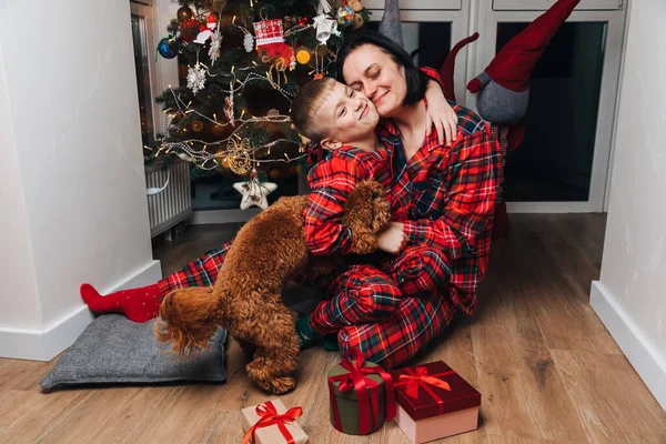 Mom and son in red pajamas and dog sit near the Christmas tree and hugging. Front view