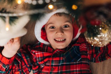 Kırmızı-yeşil pijamalı ve Noel Baba şapkalı neşeli bir çocuk yerde yatıyor. Evde Noel. Üst görünüm