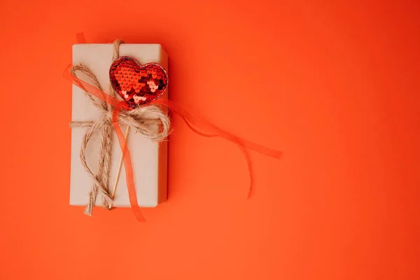 A gift wrapped in brown paper decorated with a red shiny heart on a red background. Valentines Day. Flat lay
