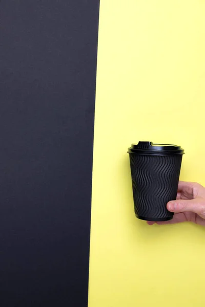 Black paper cup with coffee in a female hand on a black-yellow background. Front view