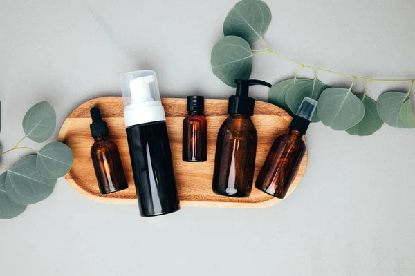 Amber bottles with facial cosmetics on a tray with a branch of eucalyptus on the gray concrete background. Top view