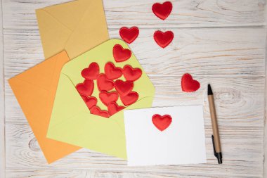 Light green, orange, yellow envelopes and a white card for text with red falling hearts on a white wooden background. Happy Valentines Day greeting, background. Flat lay