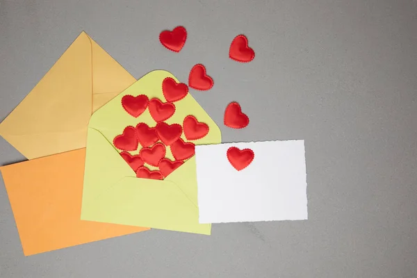 Light green, orange, yellow envelopes and a white card for text with red falling hearts on a gray concrete background. Happy Valentines Day greeting, background. Flat lay
