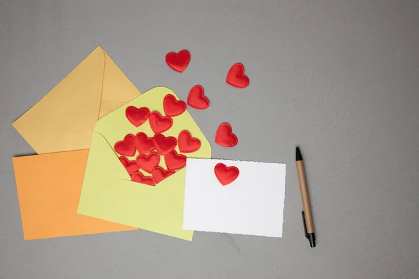 Light green, orange, yellow envelopes and a white card for text with red falling hearts on a gray concrete background. Happy Valentines Day greeting, background. Flat lay