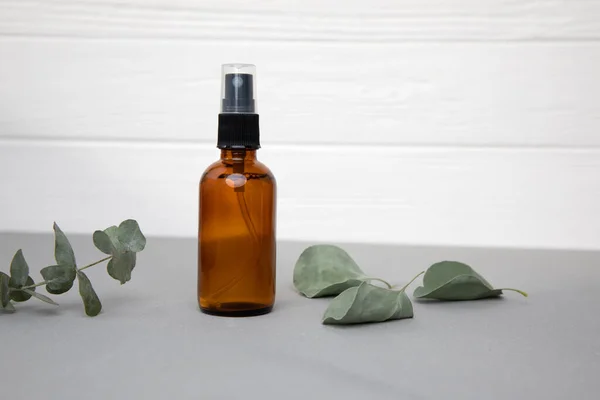 Amber spray bottle with facial cosmetics in the white-grey background with eucalyptus leaves. Front view