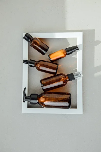 Amber bottles with facial cosmetics in the white frame on the grey concrete background. Selfcare cosmetics. Flat lay