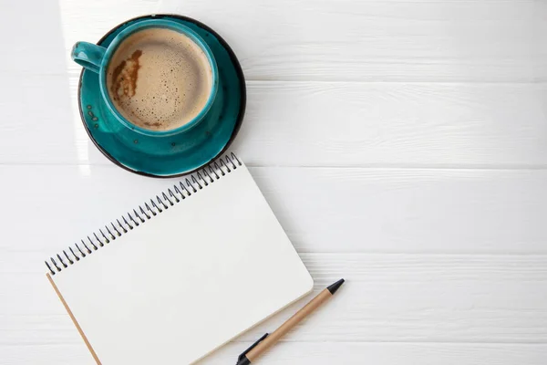 Blue mug with coffee, cappuccino with the notepad and pen on a white wooden background. Top view, close-up