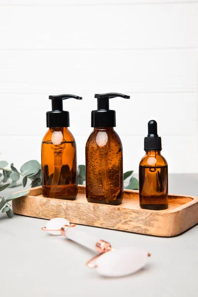 Amber bottles with facial cosmetics on a tray with a branch of eucalyptus on the grey-white background. Front view